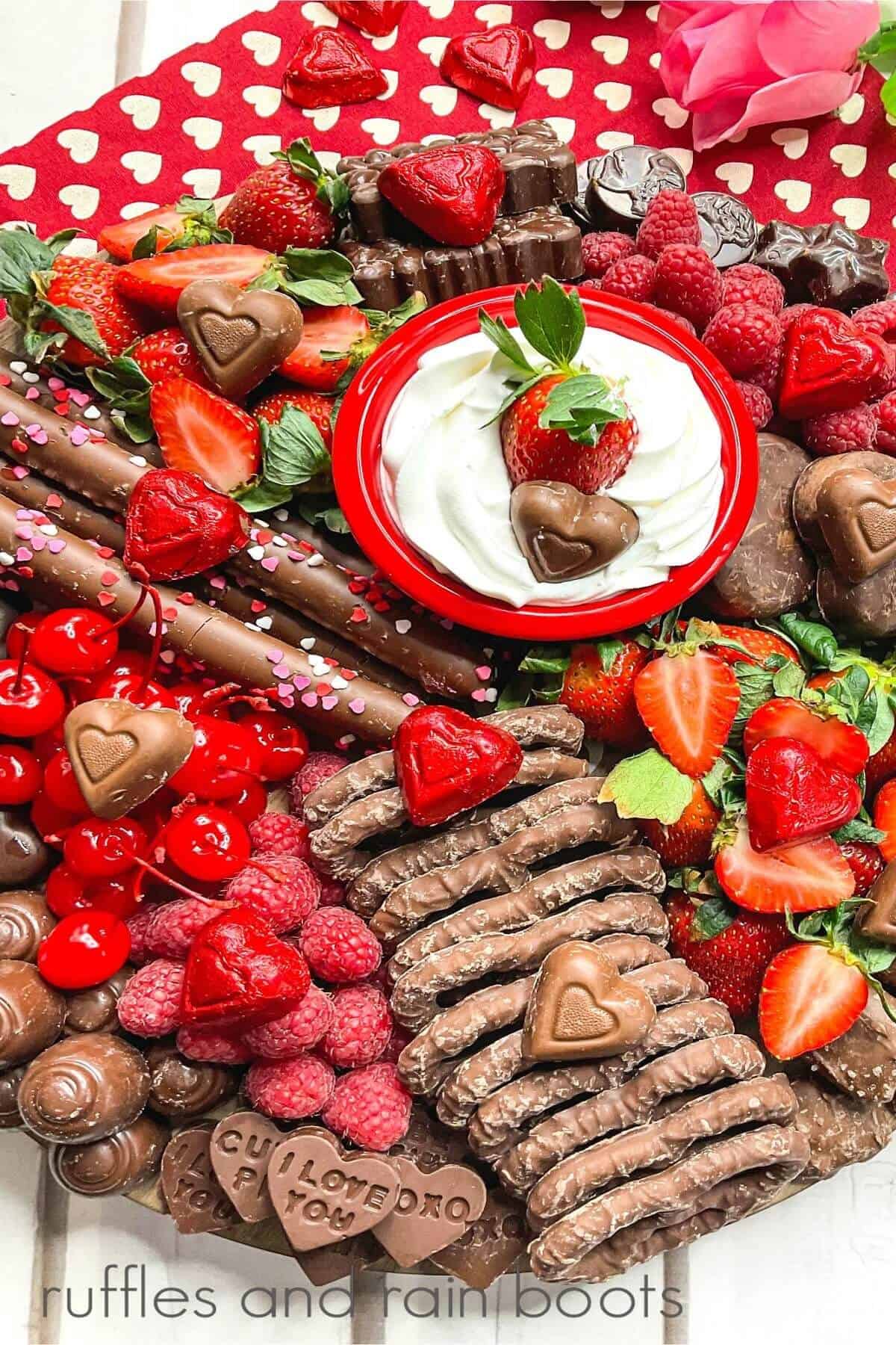 Charcuterie Board close up filled with chocolate pretzels, homemade and store-bought chocolate, fresh strawberries and raspberries, candy and a ramekin filled with dip on a red towel with white hearts on a white surface.