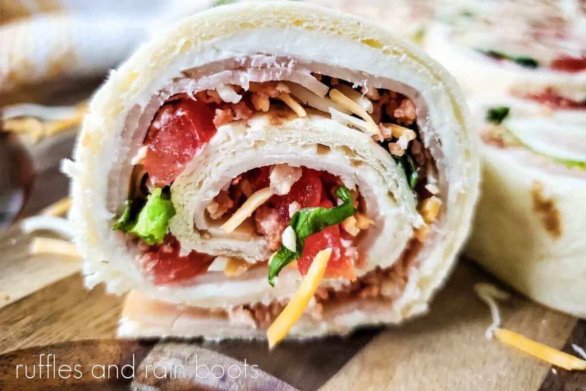 Close up of a single Overhead view of the rolled Club Sandwich Pinwheels Recipe on a wooden cutting board next to a beige and white towel.