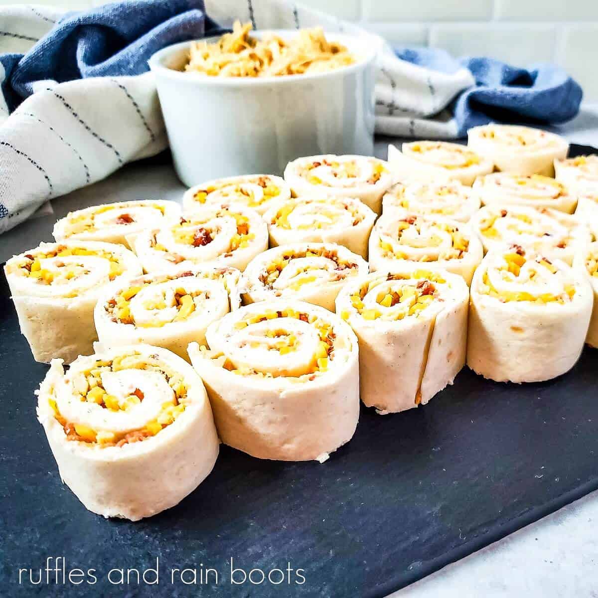 Front view image of several crack chicken pinwheel sandwiches on a black marble slab, next to a small bowl of shredded chicken and a grey and white striped towel on a white marble surface.