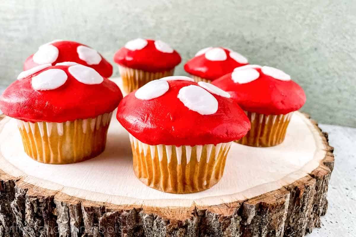 Close up horizontal image of six red and white mushroom cupcakes on a wood cutout with bark placed on concrete.