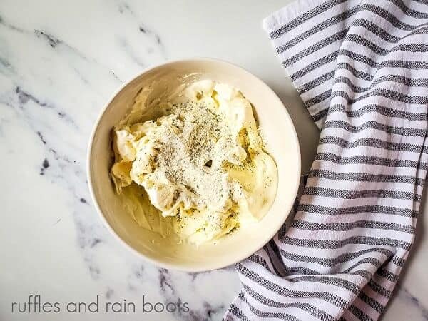 Overhead image of a small bowl of cream cheese and dry ranch dressing mix, next to a grey and white striped towel on a white marble surface.