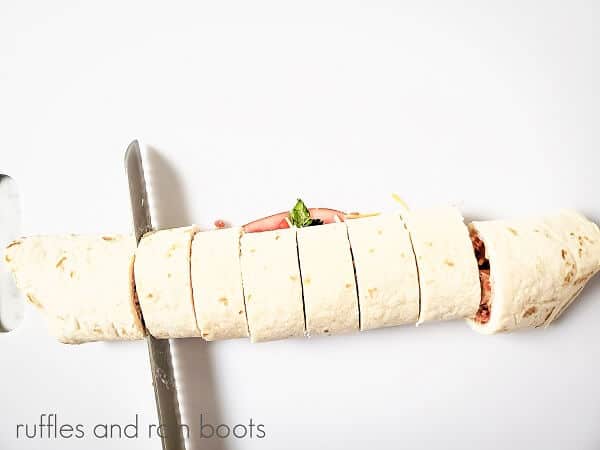 The rolled up Club Sandwich Pinwheels Recipe being cut into pieces on a white surface.