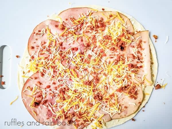 A round tortilla covered in the mayo cream cheese mixture ham, turkey, and bacon and cheese, on a white surface.