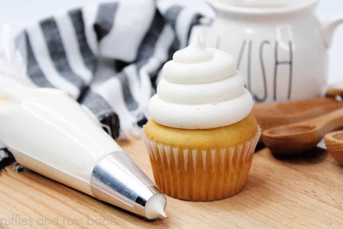 Horizontal image of round piped vanilla cupcake with buttercream frosting with piping bag, wood cutting board, and measuring spoons.