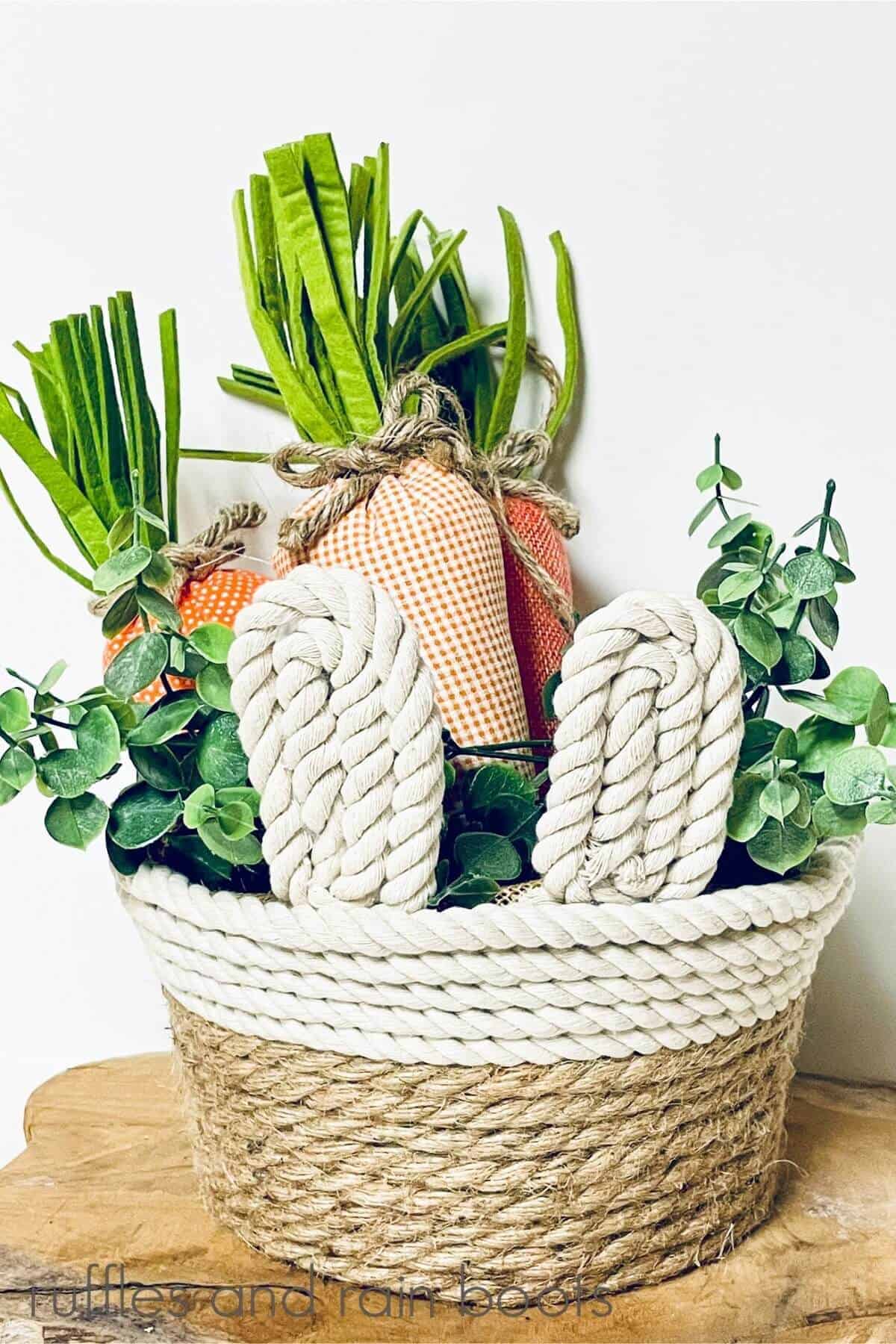 Close up of Farmhouse Rope Bunny Basket filled with faux greenery and fabric carrots with a white background on a piece of wood on a white surface.