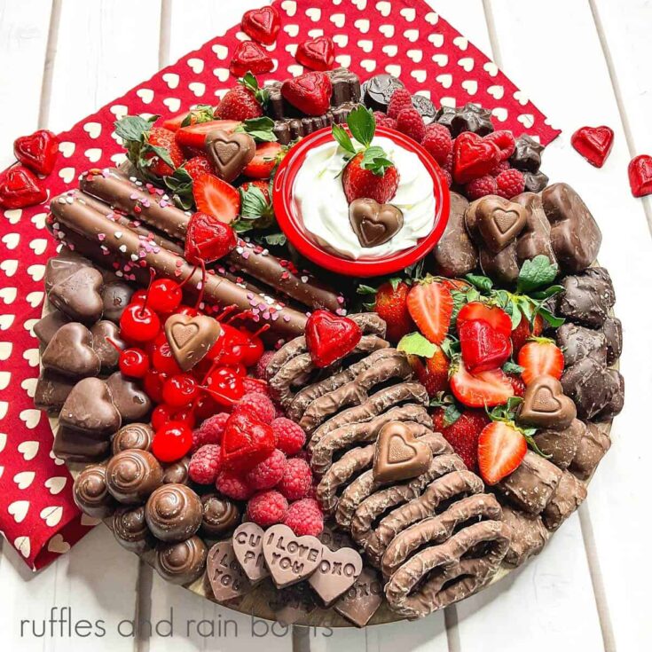Overhead view of the Chocolate Lover's Charcuterie Board filled with chocolate pretzels, various types of chocolate, berries, candy and a ramekin filled with dip on a red towel with white hearts next to faux roses on a white surface.