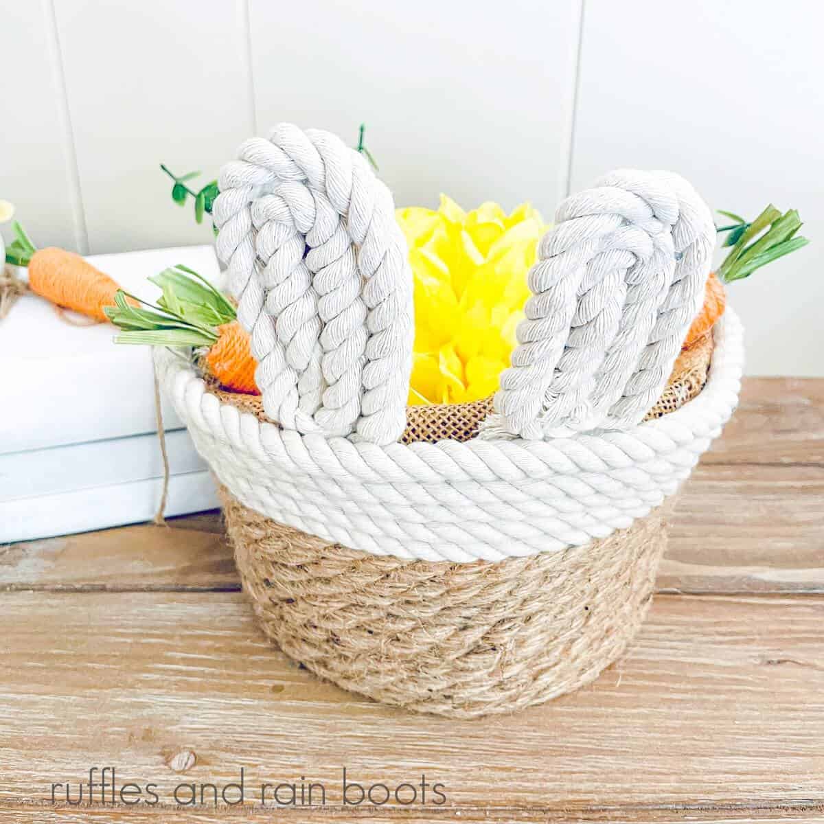 Overhead image of Farmhouse Rope Bunny Basket filled with yellow flowers and fabric carrots with a white background next to white wooden books with carrots on it on a weathered wood surface.