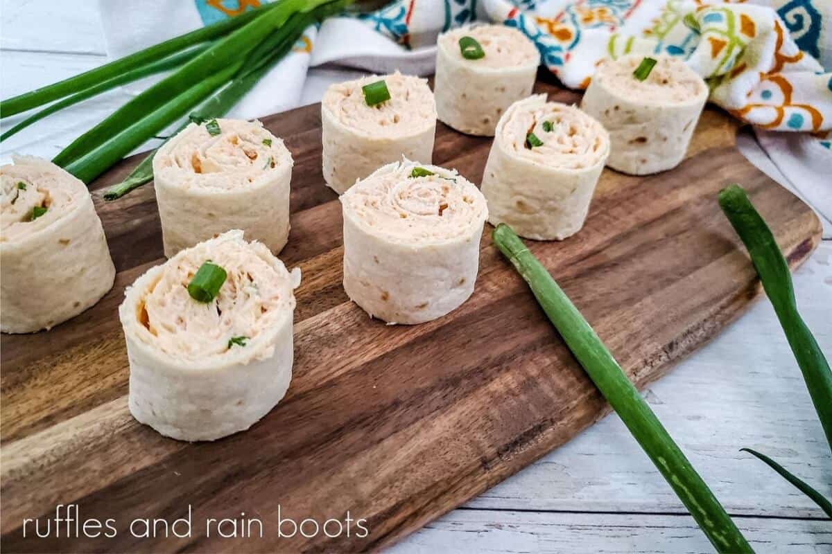 Close view of the rolled buffalo chicken pinwheel sandwiches, on a wooden cutting board, next to fresh green onion and a colorful towel on a white weathered wooden surface.