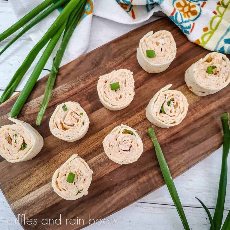 Overhead image rolled buffalo chicken pinwheel sandwiches, on a wooden cutting board, next to sprigs of fresh green onion and a colorful towel on a white weathered wooden surface.