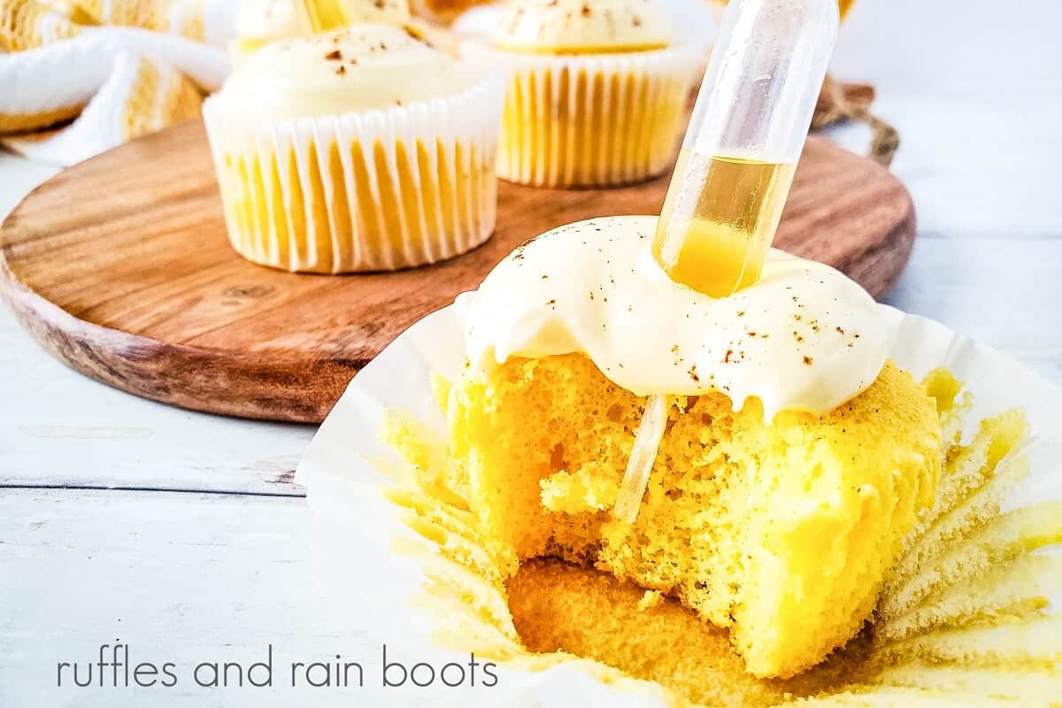 A close-up image of a cupcake, with a bite taken out, with a clear plastic pipette in the middle with a small round wooden cutting board with two cupcakes on it next to a gold and white towel on a weathered white wood surface.