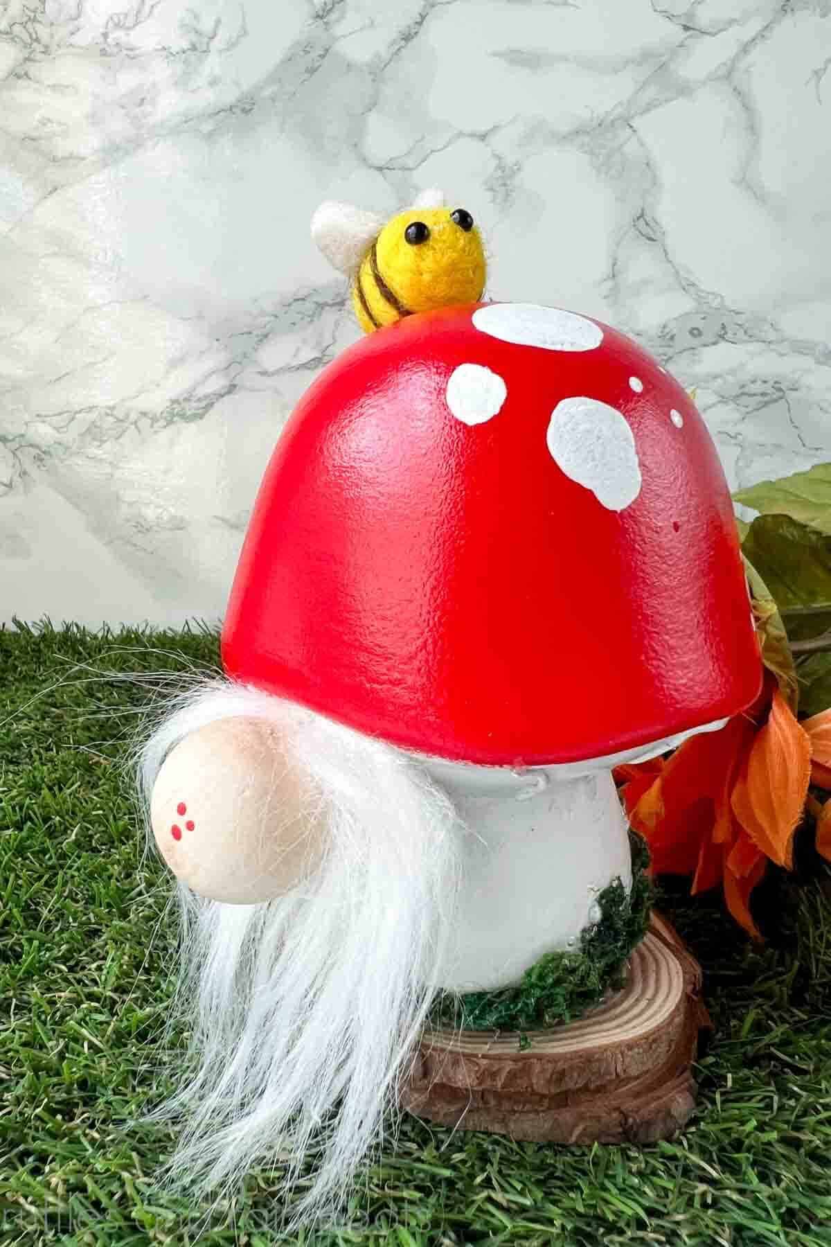 Red, white, and moss covered ceramic mushroom gnome with white beard and bee on grass and marble background.