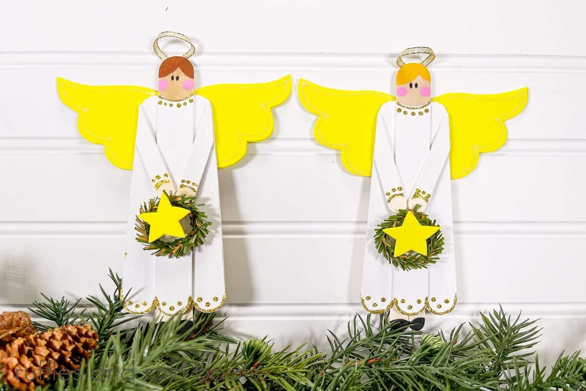 Two craft stick angels next to one another against a white wood beadboard backdrop on top of faux pine greenery and pinecones.