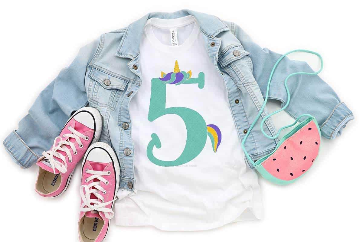 Horizontal close up of white shirt with unicorn number 5 SVG in vinyl with jean jacket, shoes, and watermelon purse.