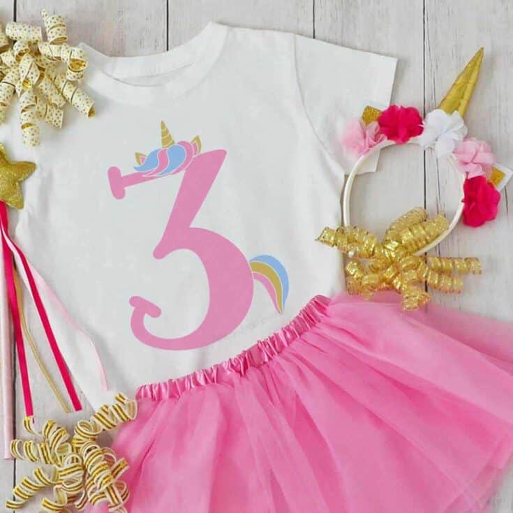 Square close up image of a white toddler shirt with unicorn number 3 in vinyl next to pink tulle skirt and unicorn headband.