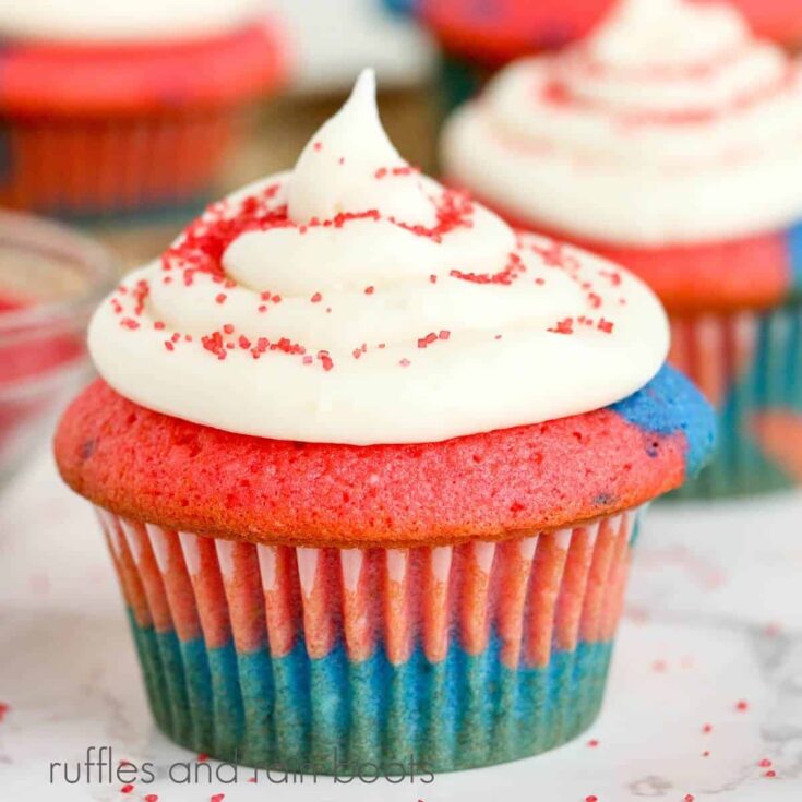 Close-up of a USA cupcake with more cupcakes in the background along with a small glass bowl of red sprinkles on a white marble background.