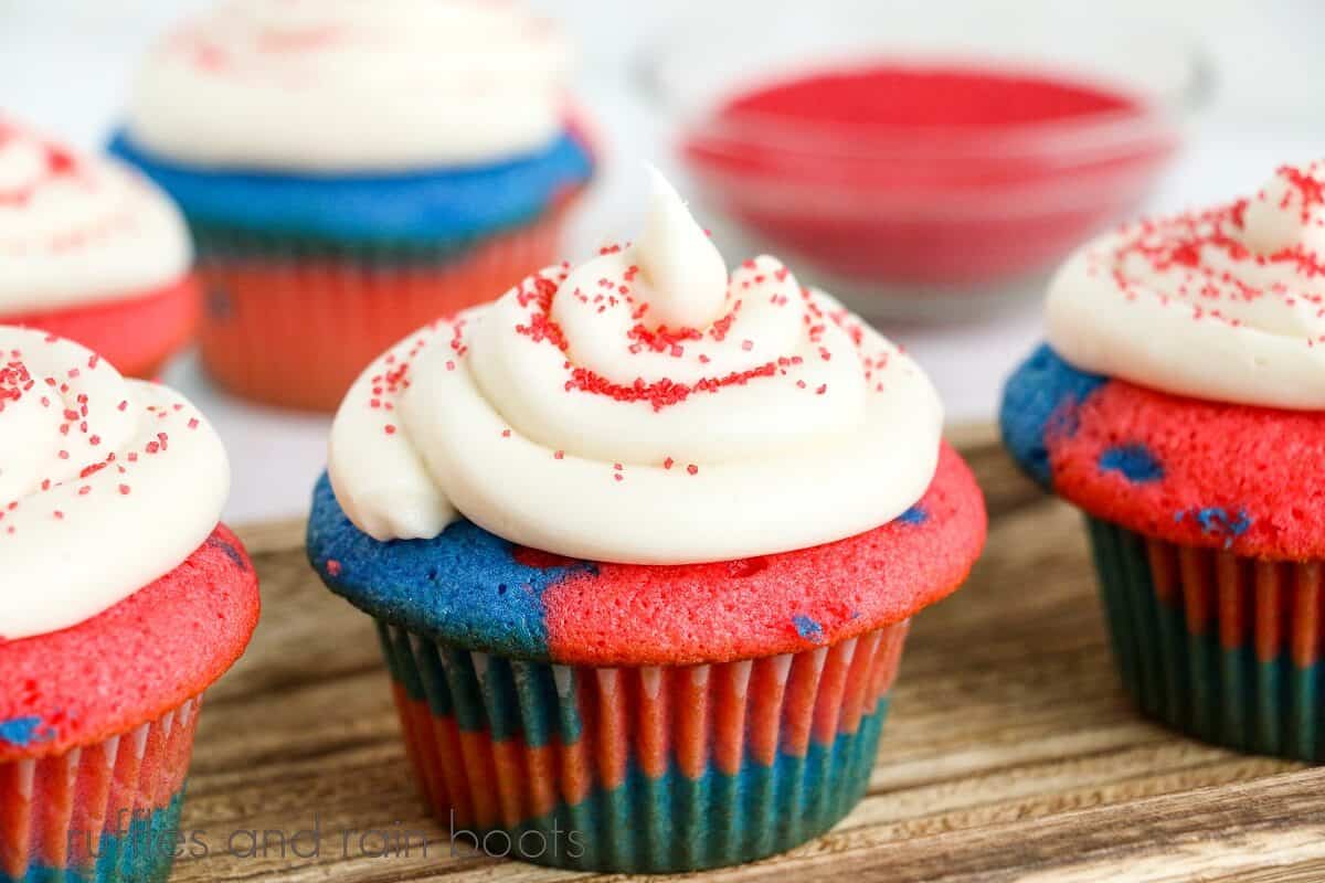 3 homemade USA cupcakes on a wooden cutting board, with 2 cupcakes in the background along with a small glass bowl of red sprinkles on a white marble background.