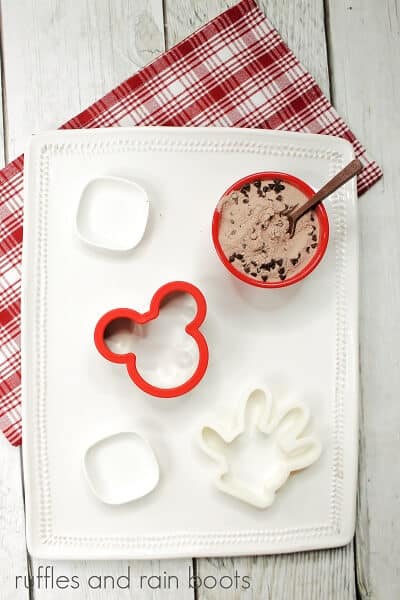 A vertical image of a white plate with Mickey's hand and Mickey's ear cookie cutter, next to a red ramekin filled with hot cocoa mix, next to a white and red plaid towel on a white wood surface.