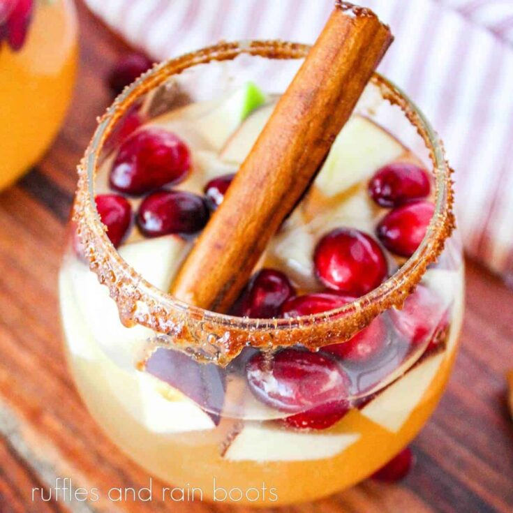 A close-up image of a two glasses filled with Sparkling Apple Cider Sangria and garnished by a cinnamon stick, on a wooden board, on a white surface.