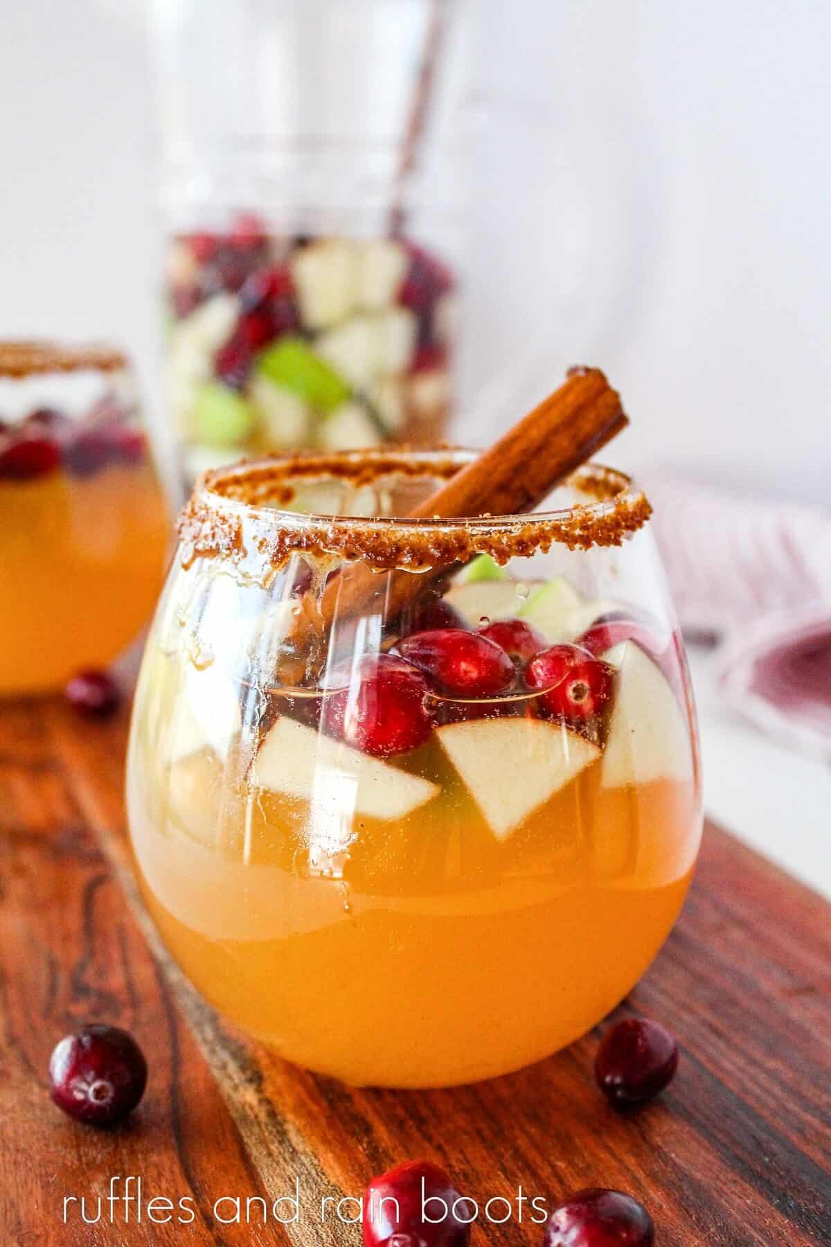 An image of a glass filled with Sparkling Apple Cider Sangria and garnished by a cinnamon stick, on a wooden board, on a white surface with another glass and a pitcher in the background.