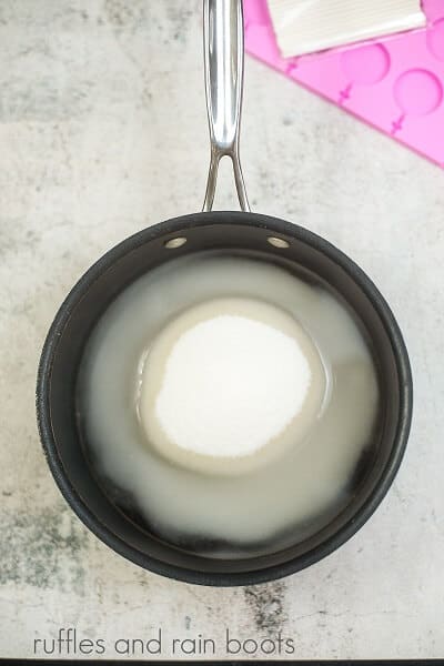 A black saucepan with water, sugar and corn syrup net to a silicone lollipop mold on a white concrete surface.