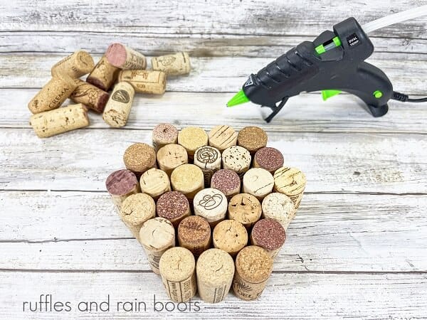 A group of recycled wine corks next to a pile of wine corks next to a hot glue gun on a white weathered wood surface.