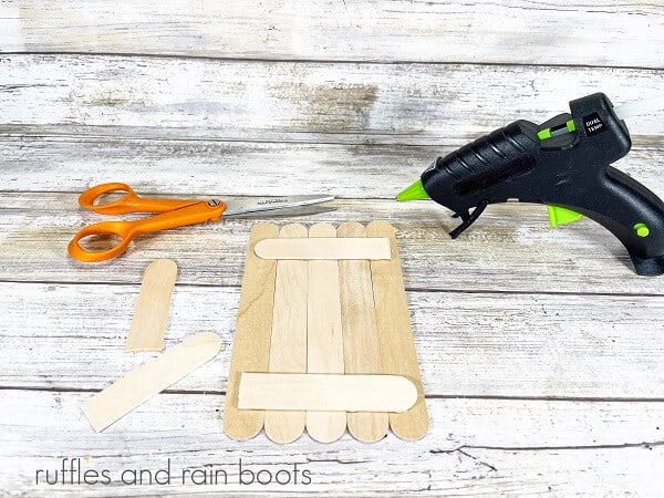 Unpainted jumbo craft sticks, with two sticks cut in half, next to a pair of orange-handled craft scissors and a hot glue gun on a white weathered wood background.