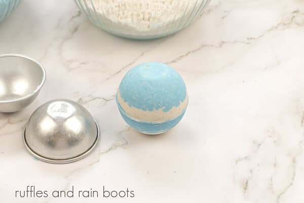 A round clear glass bowl with bath bomb mixture, next to an empty bath bomb mold, next to a large white and blue bath bomb on a white marble background.