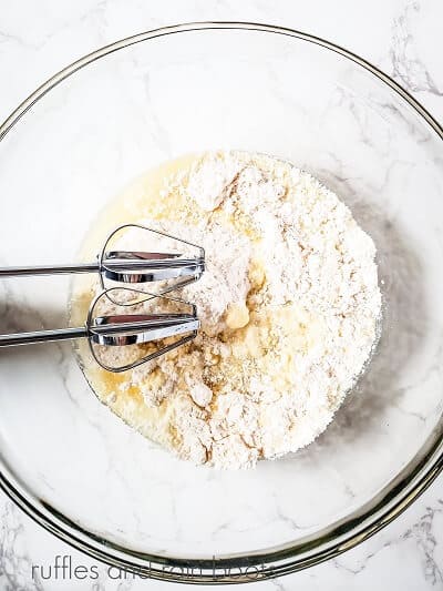 A large clear round mixing bowl with metal beaters mixing a cake mix with water and rum, against a white marble surface.