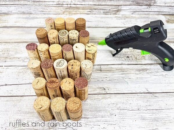 Rows of recycled wine corks next to a hot glue gun on a white weathered wood surface.