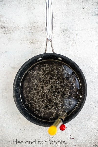 A black saucepan filled with the ingredients for the homemade lollipop recipe with a candy thermometer inside on a white concrete background.