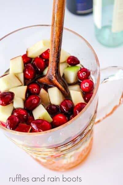 A tall glass pitcher filled wine, with fresh apples and cranberries being stirred with a wooden spoon inside on a white background.