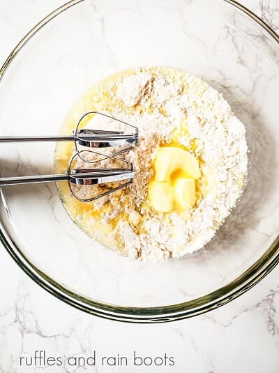 A large clear round mixing bowl with metal beaters mixing a cake mix with softened butter, against a white marble surface.