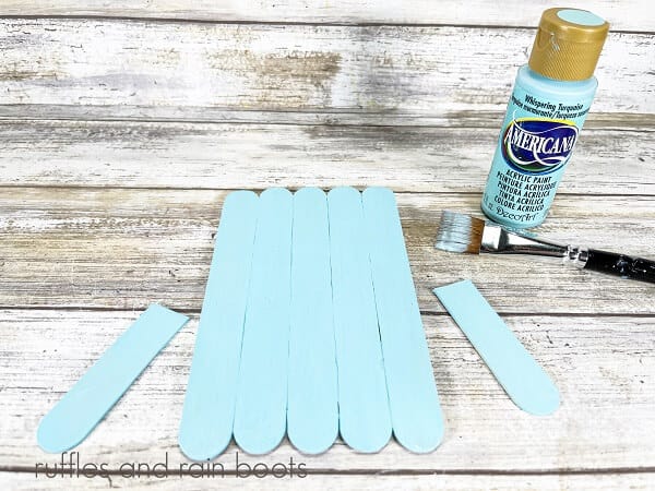 Painted jumbo craft sticks, next to a light blue bottle of craft paint and a paintbrush on a white weathered wood background.