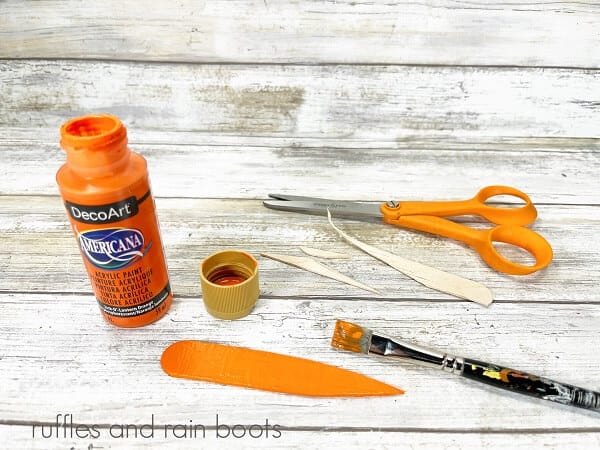 A bottle of orange acrylic paint next to a pair of scissors, a painted craft stick carrot nose and a paintbrush on a white weathered wood surface.