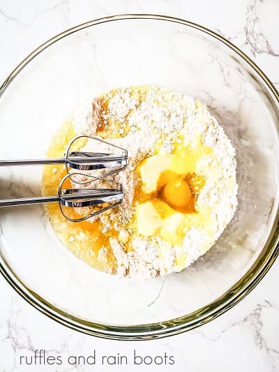 A large clear round mixing bowl with metal beaters mixing a cake mix with eggs, against a white marble surface.