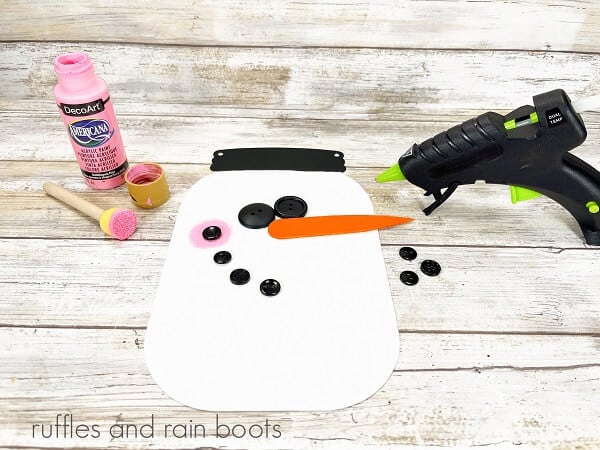 Painted Hanging Mason Jar Snowman with black button eyes, mouth, and pink cheek as well as an orange carrot nose, next to a hot glue gun and a bottle of pink acrylic paint with a foam dauber on a white weathered wood surface.