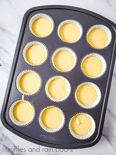 A metal cupcake pan, lined with cupcake liners and filled with batter on a white marble background.