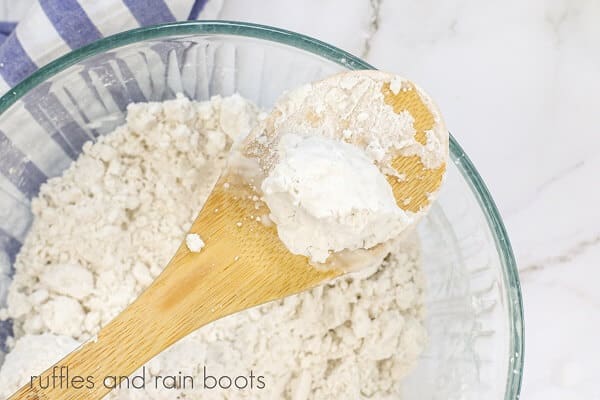 A large round glass bowl with white bath bomb mixture and a wooden spoon, next to a blue and white striped towel, on a white marble background.