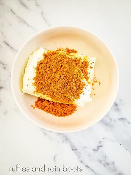 Cream cheese mixture with taco seasoning and garlic powder in a round white bowl on a white marble background.