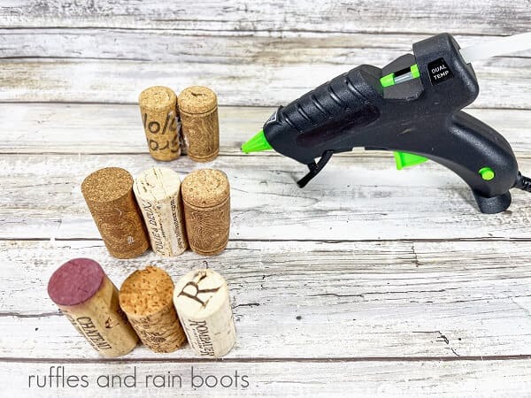 Recycled wine corks next to a hot glue gun on a white weathered wood surface.