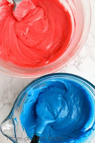 A round glass bowl of red cake batter and a bowl of blue cake batter with spoons in them on a white marble surface.