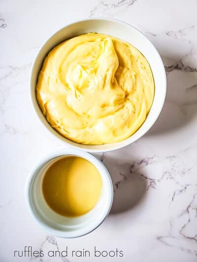 A round white bowl of buttercream frosting next to a small round white bowl of rum on a white marble surface.