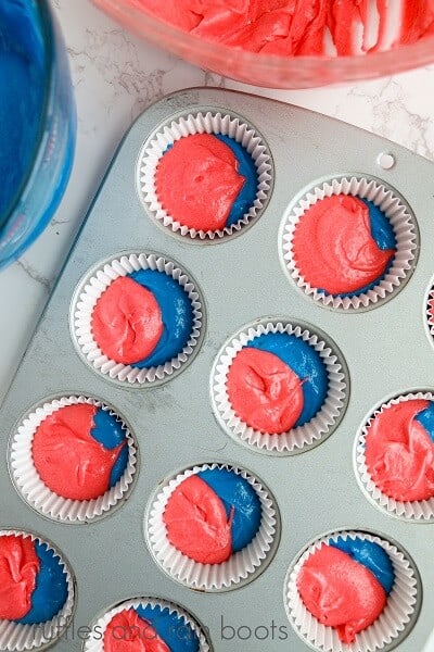 A round glass bowl of red cake batter and a bowl of blue cake batter with a cupcake pan filled with the red and blue batter on a white marble surface.