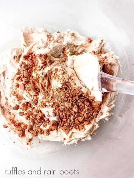 A small round glass bowl filled with frosting and chopped butterscotch chips and a spatula on a white surface