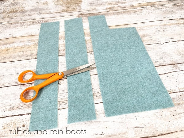 3 cut scraps of blue felt next to a pair of orange-handled scissors on a white weathered wood surface.