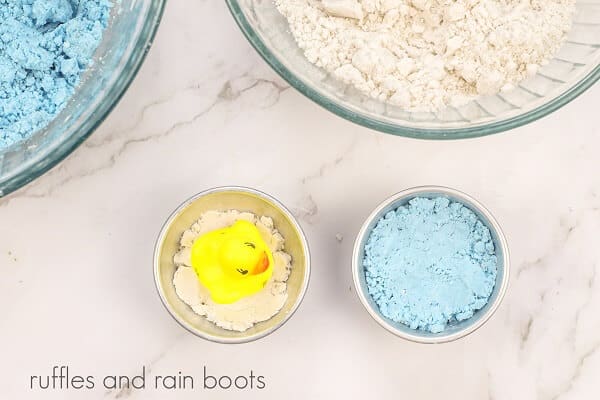 Two round glass bowls, one with blue and one with white bath bomb mixture, next to a small duck in one side of the bath bomb mold and mixture in the other side of the mold.