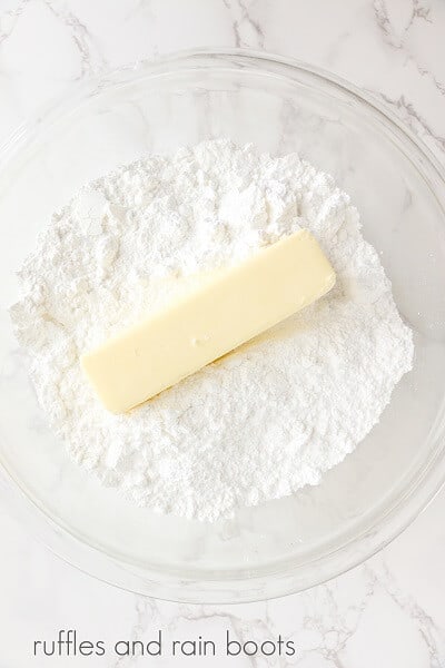 A round glass bowl with powdered sugar and a stick of butter on a white marble surface.