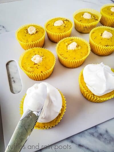 Several banana cream pie cupcakes, filled with pudding, with one being frosted on a white cutting board, on a white marble surface.
