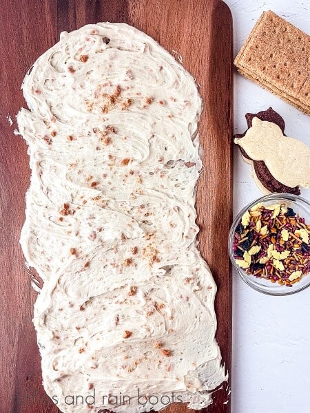 An image of a rectangle cutting board with Butterbeer frosting on the top, with a small bowl of sprinkles and cookies and cracker next to it on a white surface.