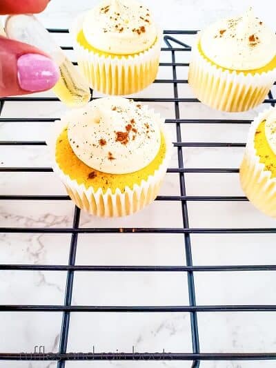 A hand placing a pipette filled with rum into a cupcake that is frosted and sprinkled with cinnamon, on a wire rack with 3 other cupcakes on a white marble surface.